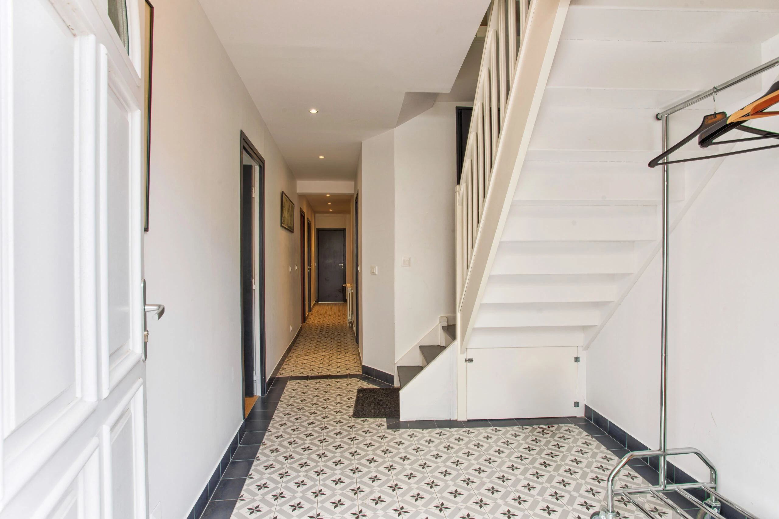 Entrée de la maison avec escalier et carrelage géométrique – maison Laguna Entrée avec escalier blanc, carrelage noir et blanc à motifs, et penderie ouverte – maison Laguna