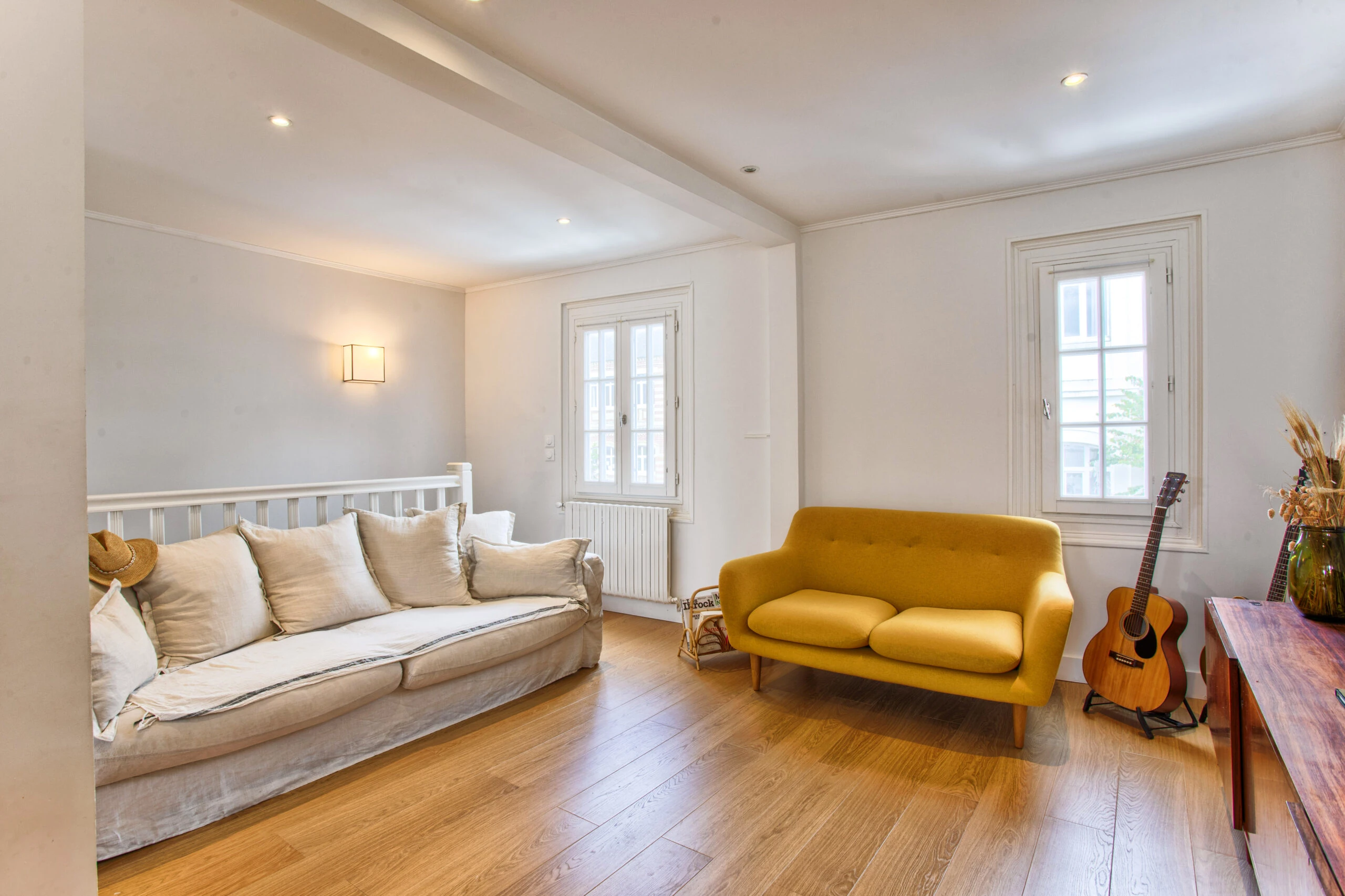 Salon lumineux avec canapé jaune et guitare – maison Laguna Salon cosy avec canapé jaune, fauteuil blanc, guitare et grandes fenêtres – maison Laguna