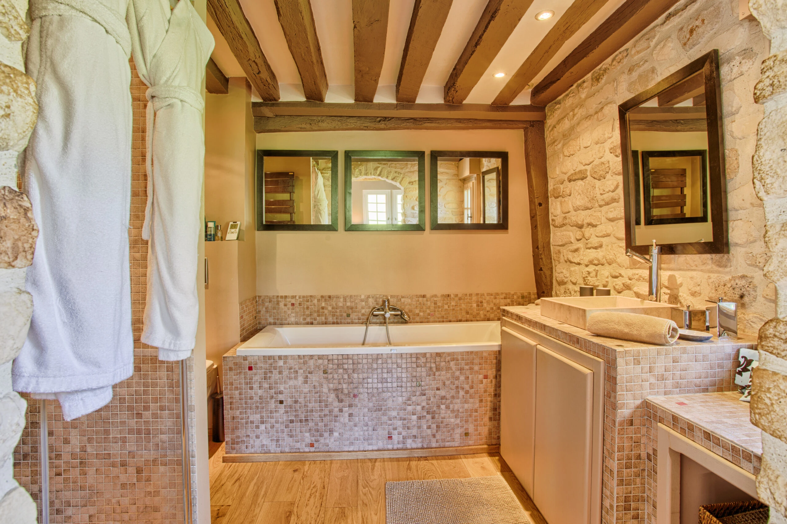 Salle de bain en pierre avec grande baignoire – Villa Constantin Salle de bain élégante avec murs en pierre, baignoire encastrée et trois miroirs dans la Villa Constantin