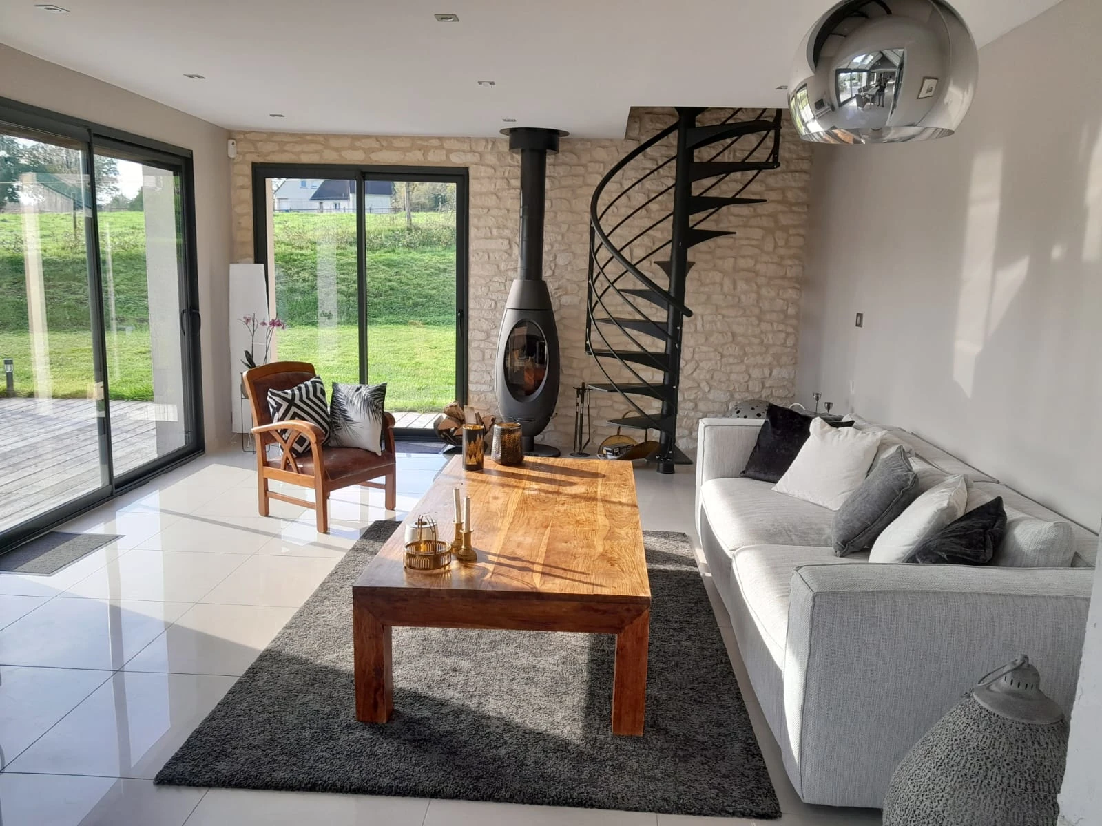 Salon lumineux avec escalier en colimaçon – Les Glycines Pont l’Évêque Salon élégant avec canapé clair, table en bois massif et escalier en colimaçon au fond, dans le logement Les Glycines.